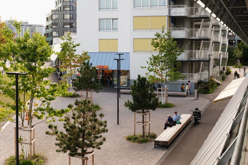 Areal Guggach – Der neue Quartierplatz an der Tramhaltestelle macht vielfältige Angebote über das Areal hinaus. Hier trifft man sich zum Einkaufen, geht zum Velomech, trinkt einen Kafi oder begegnet sich beiläufig auf dem Nachhauseweg. Mit jedem Jahr wachsen die Baumkronen und spenden Schatten an heissen Sommertagen. – Max Creasy
